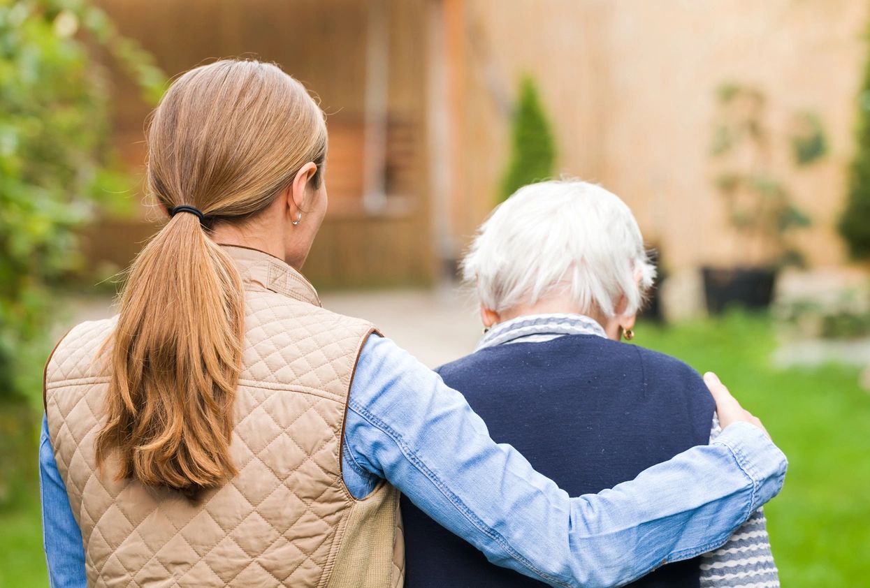 A caregiver walking outdoors with a resident, arm around her shoulder